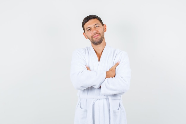 Young male standing with crossed arms in white bathrobe and looking glad , front view.