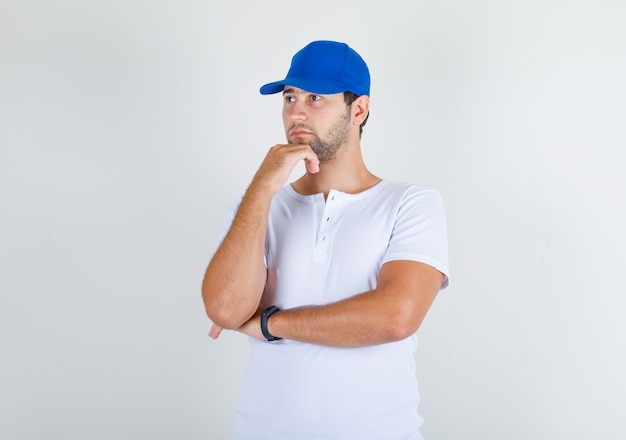 Young male standing with hand on chin in white t-shirt and blue cap