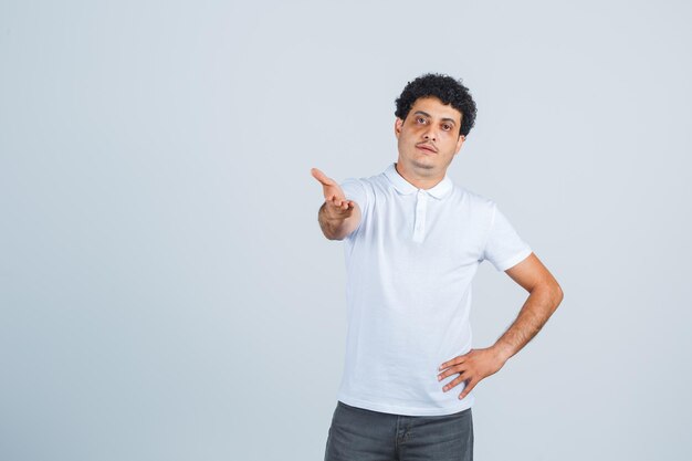 Young male stretching hand in puzzled gesture in white t-shirt, pants and looking confident. front view.