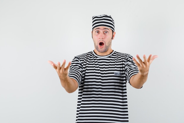 Young male stretching hands in questioning gesture in t-shirt, hat and looking surprised. front view.