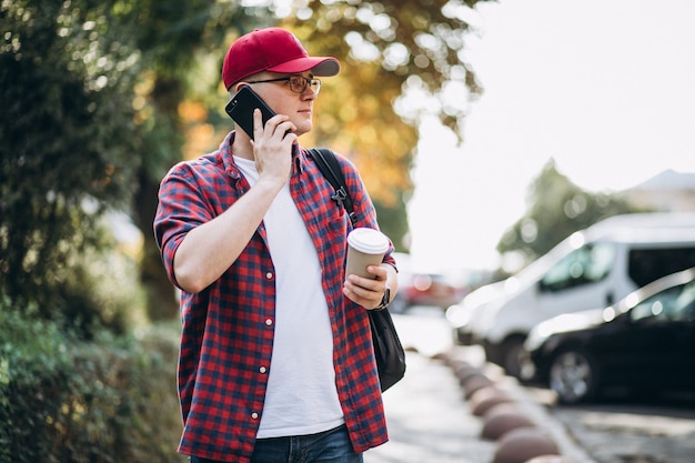 Free photo young male student drinking coffee using phone in park