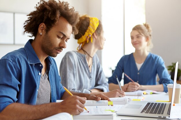 Young male student with Afro hairstyle, reading book attentively, sitting at front of opened laptop in classroom and his two female groupmates chatting with each other. Education and teamwork concept