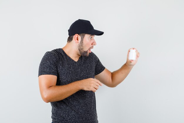 Young male in t-shirt and cap looking at pill bottle and looking surprised