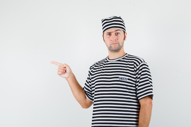 Young male in t-shirt, hat pointing to the left side and looking hesitant , front view.