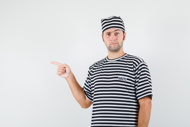 Young male in t-shirt, hat pointing to the left side and looking hesitant , front view.