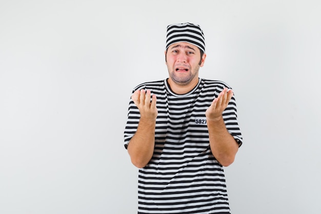 Young male in t-shirt, hat praying while crying and looking helpless , front view.