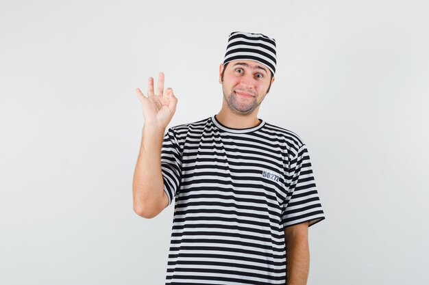 Young male in t-shirt, hat showing ok gesture and looking cheerful , front view.