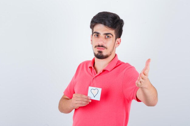 Young male in t-shirt holding sticky note while raising hand in questioning manner and looking confident , front view.