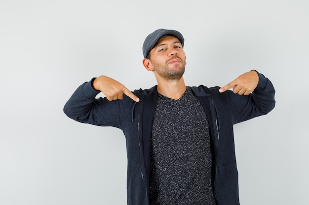 Young male in t-shirt, jacket, cap pointing at himself and looking confident , front view.