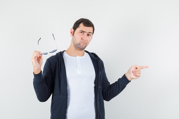 Young male in t-shirt, jacket holding glasses, pointing aside and looking hesitant , front view.