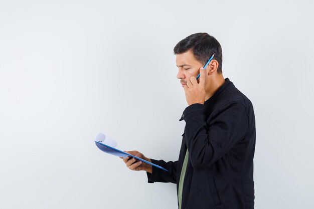 Young male in t-shirt, jacket looking over clipboard while holding pencil and looking pensive .