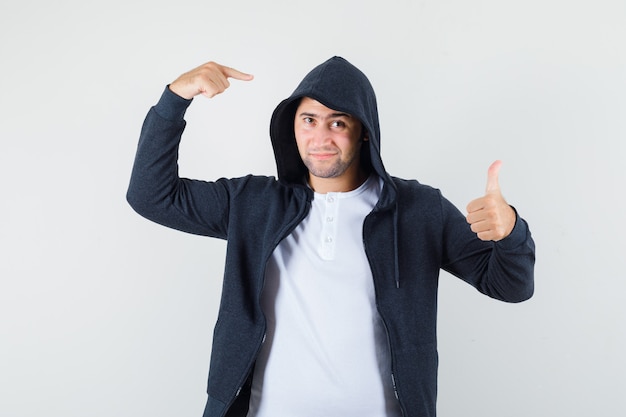 Free Photo young male in t-shirt, jacket pointing at his hood, showing thumb up and looking confident , front view.