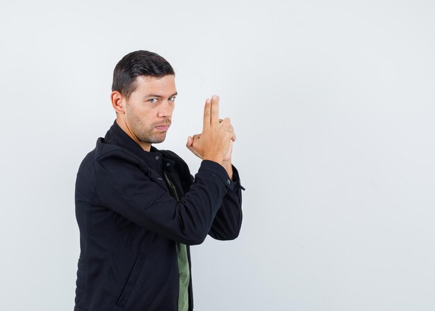 Young male in t-shirt, jacket showing gun gesture and looking serious , front view.