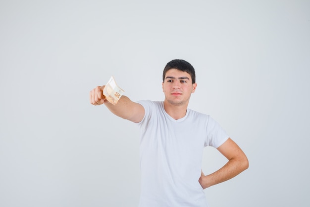 Young male in t-shirt stretching out a hand with euro banknote and looking confident , front view.