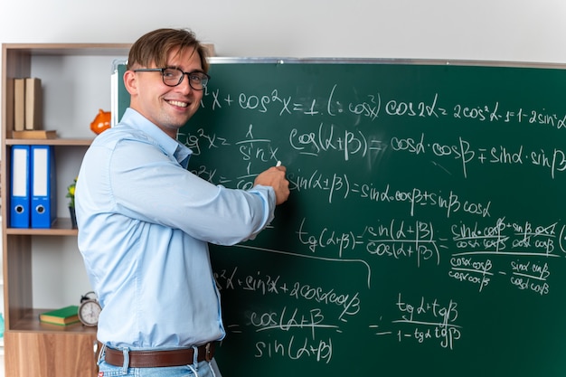 Young male teacher wearing glasses explaining lesson happy and positive smiling standing near blackboard with mathematical formulas in classroom