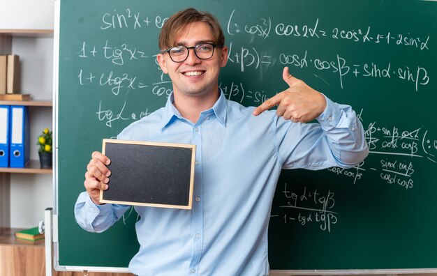 Young male teacher wearing glasses holding small blackboard pointing with index finger at it looking smiling confident standing near blackboard with mathematical formulas in classroom