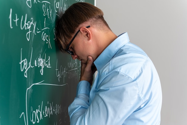 Young male teacher wearing glasses looking puzzled having no answer standing near blackboard with mathematical formulas in classroom