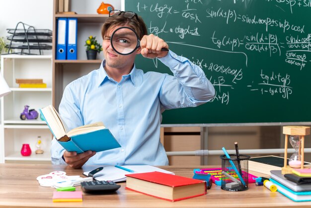 Young male teacher wearing glasses looking through magnifying glass sitting at school desk with books and notes in front of blackboard in classroom