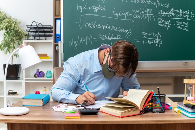 Young male teacher wearing glasses and protective facial mask writing preparing lesson sitting at school desk with books and notes in front of blackboard in classroom