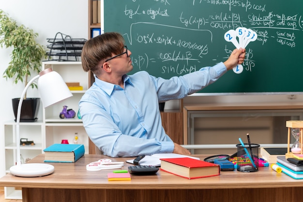 Young male teacher wearing glasses showing number plates explaining lesson smiling confident sitting at school desk with books and notes in front of blackboard in classroom