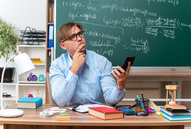 Young male teacher wearing glasses typing message using smartphone looking puzzled sitting at school desk with books and notes in front of blackboard in classroom