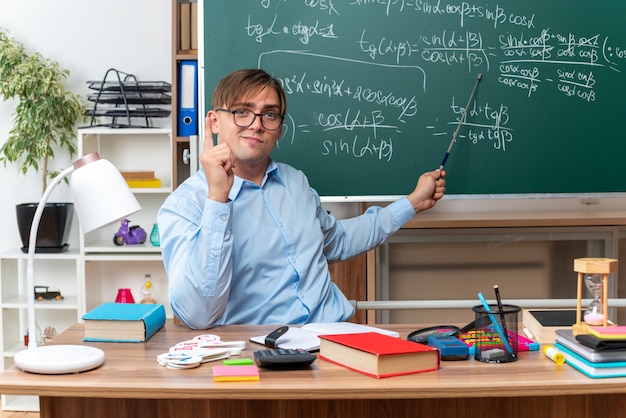 Young male teacher wearing glasses with pointer explaining lesson looking confident sitting at school desk with books and notes in front of blackboard in classroom