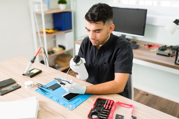 Free photo young male technician using compressed air to clean inside a dirty smartphone at the repair shop. latin man with gloves cleaning a cellphone