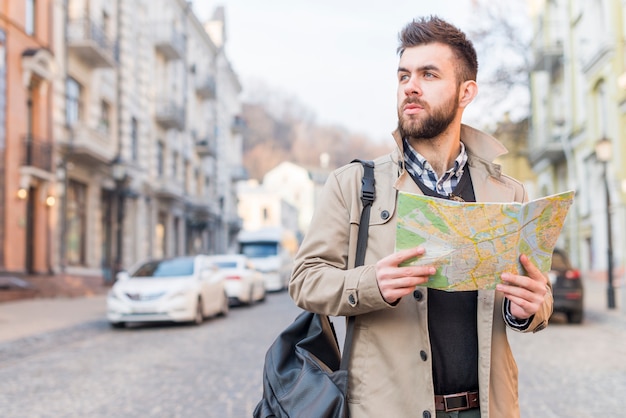 Young male tourist with bag on his shoulder standing on street holding map in hand looking away