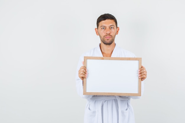 Free photo young male in white bathrobe holding white board and looking strict , front view.