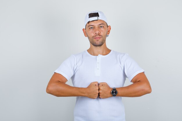 Young male in white t-shirt, cap bumping his fists and looking confident, front view.