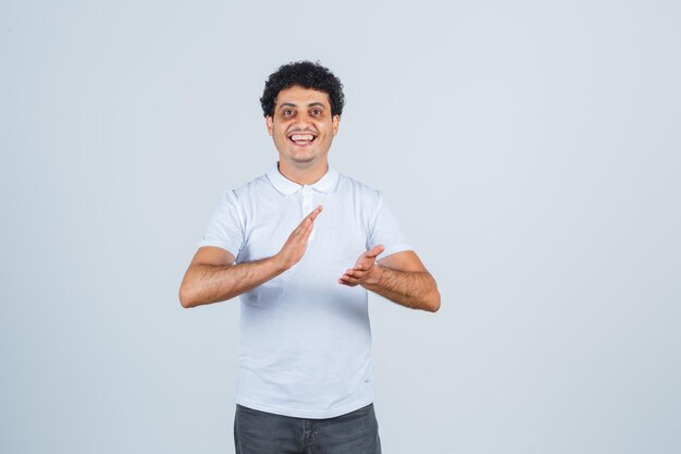 Young male in white t-shirt, pants clapping hands after great performance and looking merry , front view.