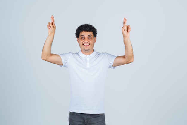 Young male in white t-shirt, pants keeping fingers crossed and looking happy , front view.