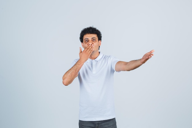 Young male in white t-shirt, pants keeping hand on mouth, showing something and looking elegant , front view.