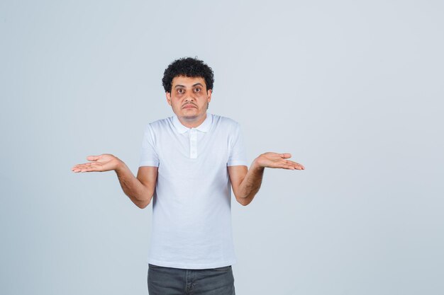 Young male in white t-shirt, pants showing helpless gesture and looking confused , front view.