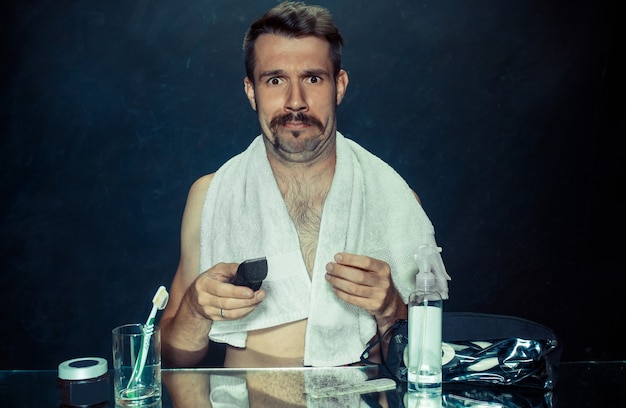 Free Photo the young man in bedroom sitting in front of the mirror scratching his beard at home. human emotions concept and problems with skin