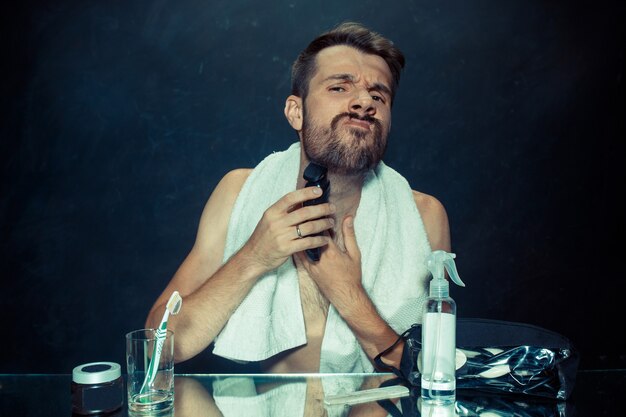 The young man in bedroom sitting in front of the mirror scratching his beard at home. Human emotions concept