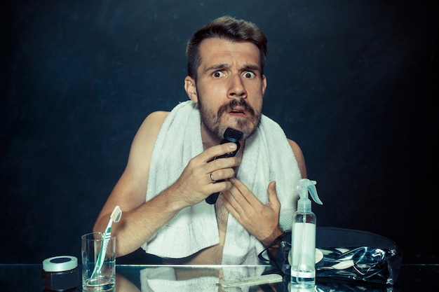 Free photo the young man in bedroom sitting in front of the mirror scratching his beard at home. human emotions and lifestyle concept