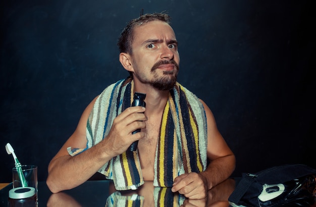 Free photo the young man in bedroom sitting in front of the mirror scratching his beard at home. human emotions and lifestyle concept