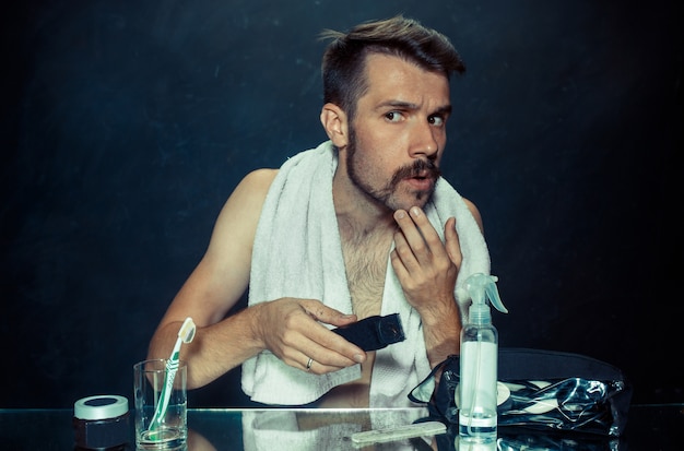 Free photo young man in bedroom sitting in front of the mirror scratching his beard at home. human emotions and lifestyle concept