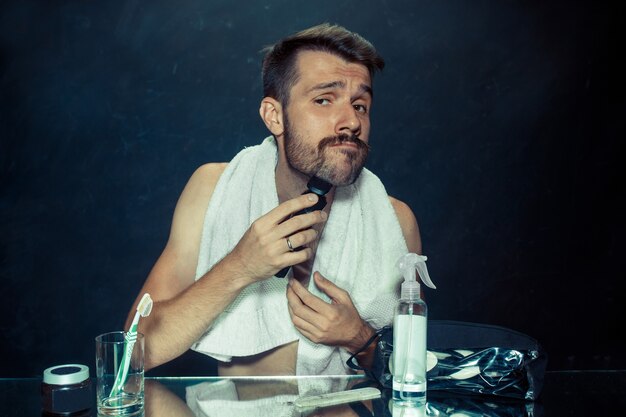 The young man in bedroom sitting in front of the mirror scratching his beard at home. Human emotions and lifestyle concept