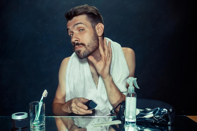 Free photo the young man in bedroom sitting in front of the mirror scratching his beard at home. human emotions and lifestyle concept