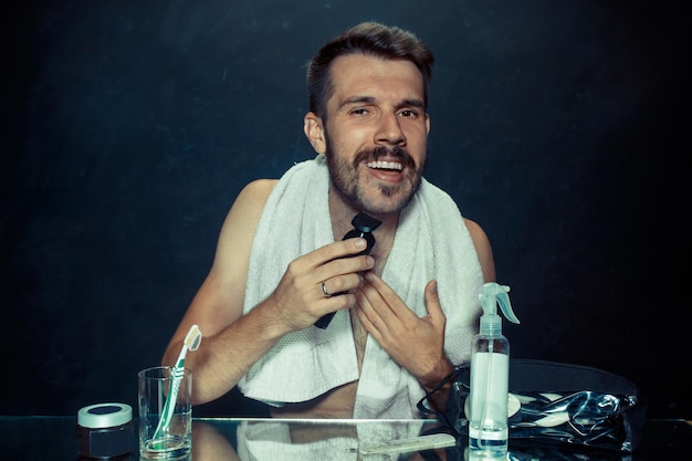 Free Photo the young man in bedroom sitting in front of the mirror scratching his beard at home. human emotions and lifestyle concept