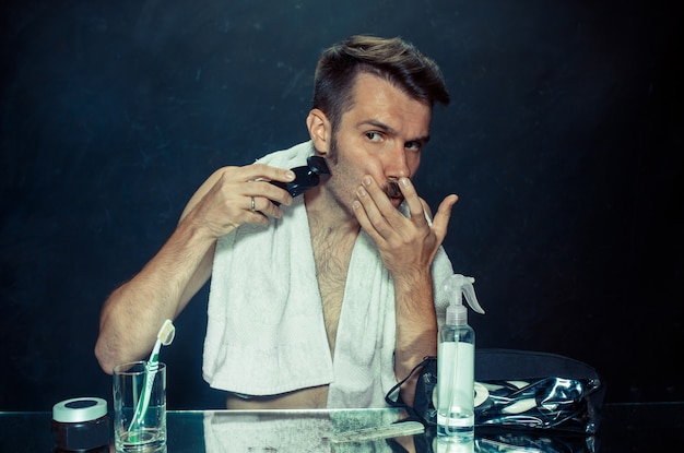 Free photo young man in bedroom sitting in front of mirror scratching his beard at home