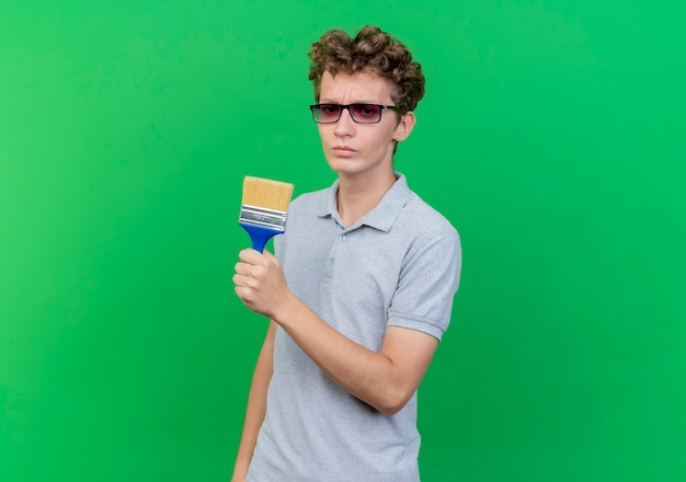 Young man in black glasses wearing grey polo shirt holding paint brush loking at camera with serious face over green