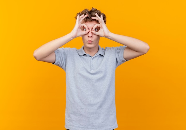 Young man in black glasses wearing grey polo shirt making binocular gesture with fingers looking through fingers over orange