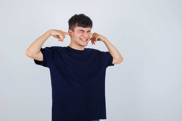 Young man in black t-shirt plugging ears with fingers and looking annoyed , front view.