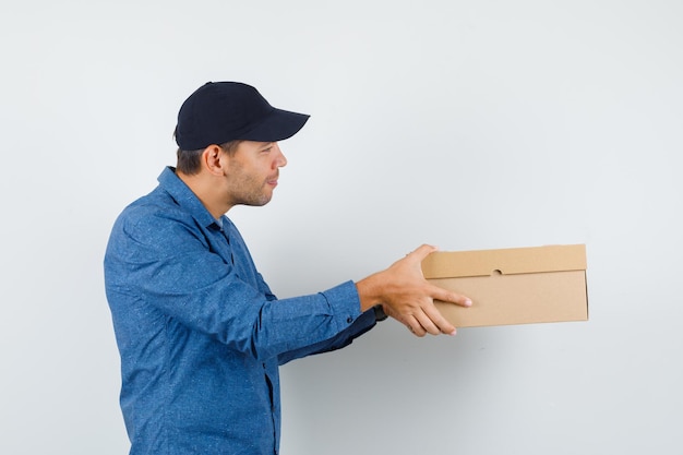 Young man in blue shirt, cap delivering cardboard box and looking cheerful .