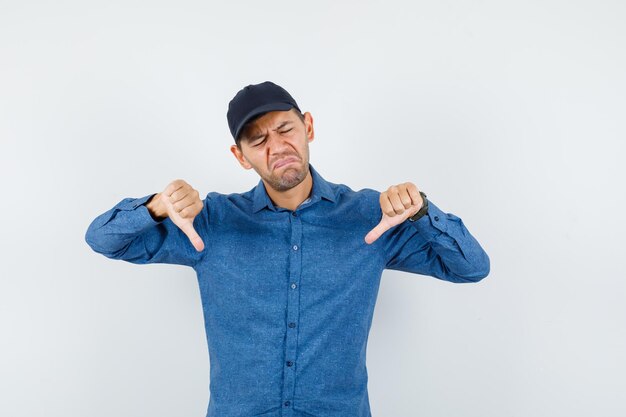 Young man in blue shirt, cap showing thumbs down and looking disappointed , front view.