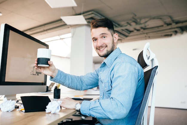 Young man in blue shirt is sitting at his workplace in office. He wears blue shirt.  He holds a cup and smiles to the camera.