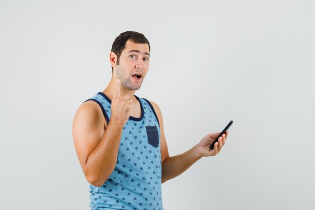 Young man in blue singlet holding mobile phone, pointing up and looking curious